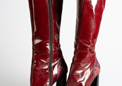 Ein Paar rote Lackstiefel steht auf einem weißen Hintergrund. Die Stiefel sind aus glänzendem rotem Material und haben einen rechteckigen Absatz. Sie sind mit einer Metallzippertasche versehen, die sich über den gesamten Fuß erstreckt. Der Hintergrund ist weiß und bietet eine klare Darstellung der Stiefel.