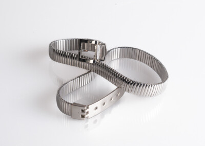 Das Bild zeigt eine silberfarbene, flexible Kette. Die Kette ist aus dünnen, miteinander verbundenen Metallstreifen gefertigt und biegt sich leicht. Sie hat einen silbernen Verschluss mit einer kleinen Schnalle. Das Licht reflektiert stark von der Oberfläche der Kette.