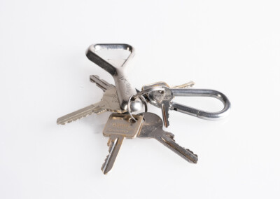 Ein Foto zeigt eine Sammlung von Schlüsseln, die an einem silberfarbenen Schlüsselanhänger aufgehängt sind. Die Schlüssel scheinen aus Metall zu bestehen und haben unterschiedliche Formen und Größen. Der Hintergrund ist weiß und glatt.