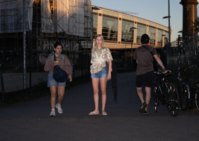 Die Aufnahme zeigt einen belebten Bahnsteig in Berlin. Eine Gruppe von Studenten der Ostkreuzschule für Fotografie spaziert am Abend entlang, während sich im Hintergrund ein großer Bahnhof befindet. Die Szene ist von einer warmen, goldenen Beleuchtung geprägt, die durch das Fenster des Bahnhofs dringt.