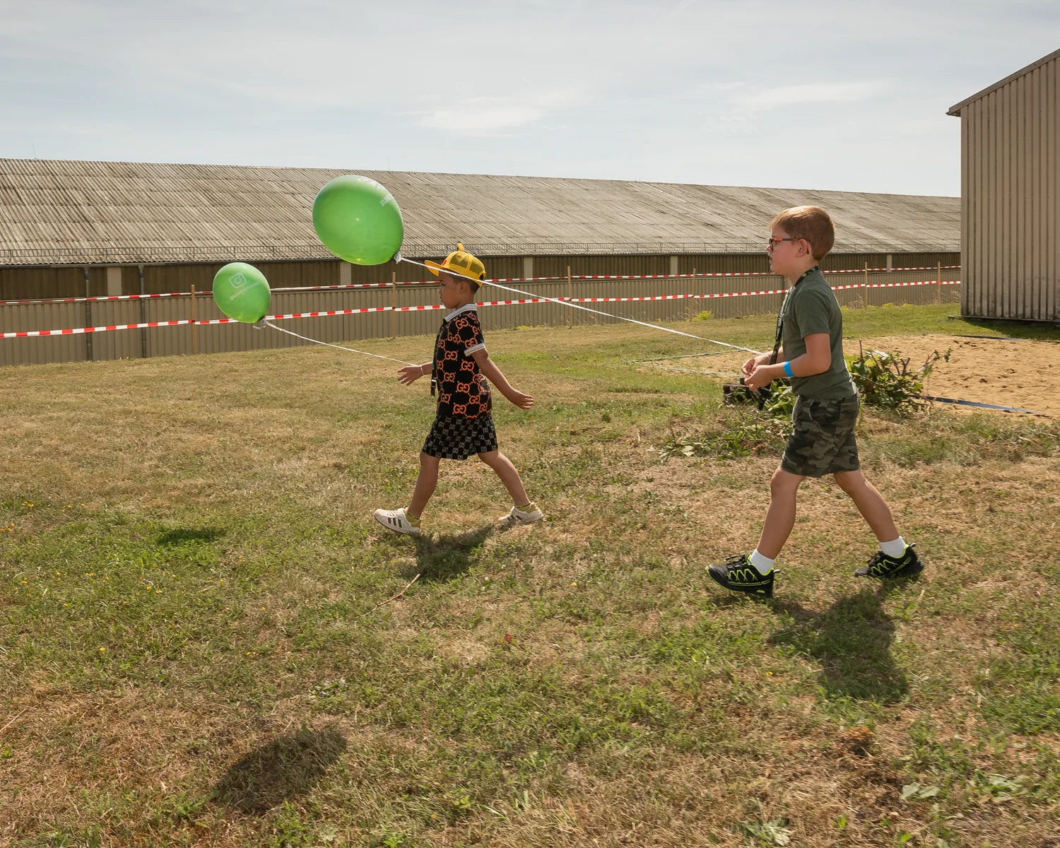 Das Bild zeigt zwei junge Männer, vermutlich Kinder, die auf einer grünen Wiese spielen. Ein Junge trägt eine gelbe Mütze und einen schwarzen T-Shirt mit einem Gucci-Motiv. Er versucht, einen grünen Luftballon zu fangen. Der andere Junge trägt eine khaki-farbene Hose und blaue Schuhe. Beide Kinder scheinen Spaß zu haben. Im Hintergrund befindet sich ein Gebäude mit einem Schieferdach. Die Sonne scheint und der Himmel ist leicht bewölkt.