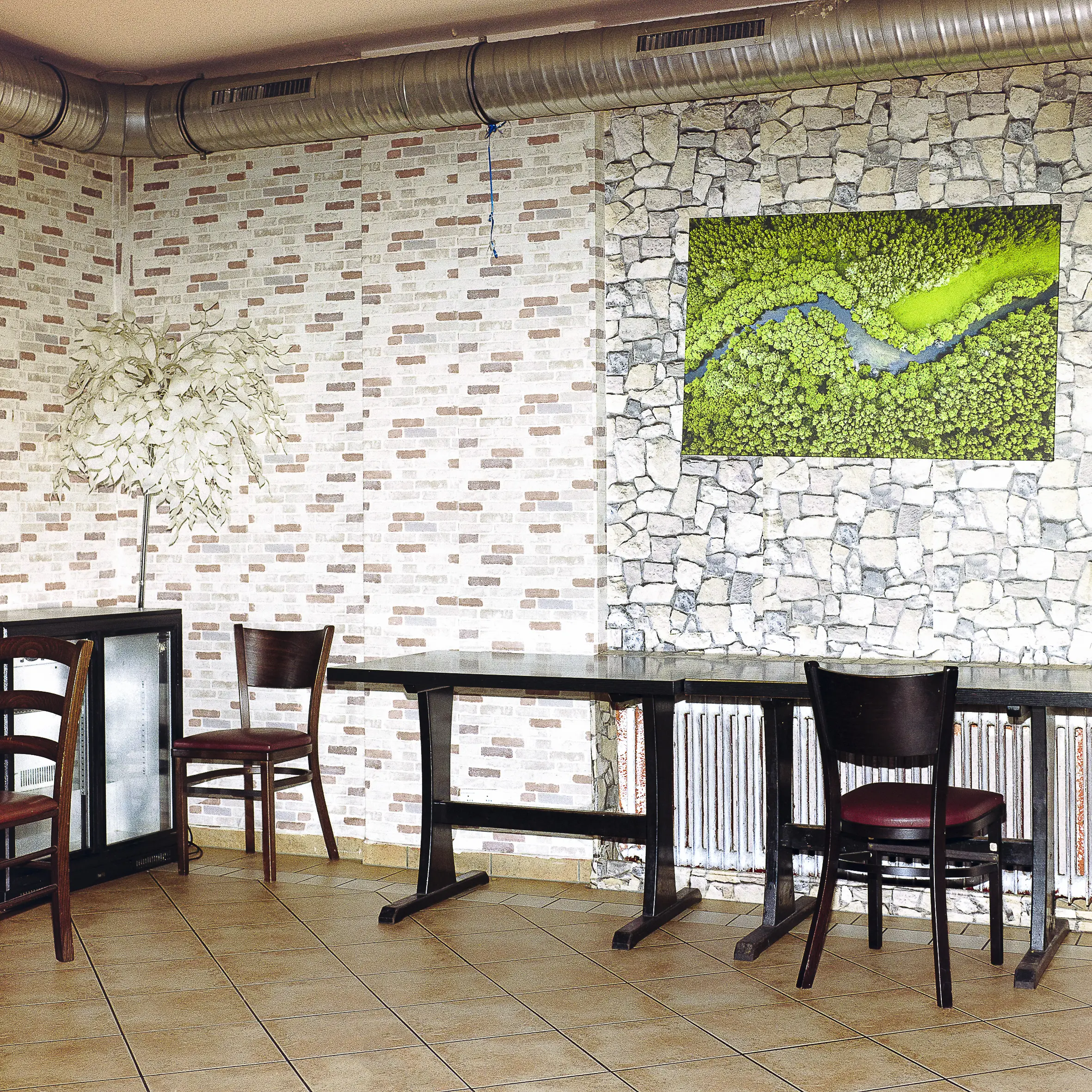 Das Bild zeigt einen Innenraum, der wahrscheinlich ein Restaurant oder eine Bar ist. Der Raum ist durch die Verwendung verschiedener Materialien wie Ziegel, Stein und Holz gestaltet. Die Einrichtung besteht aus mehreren Tischen mit Stühlen, sowie einigen Kunstwerken an den Wänden. Es gibt auch einige technische Elemente wie Rohre und Heizkörper.
