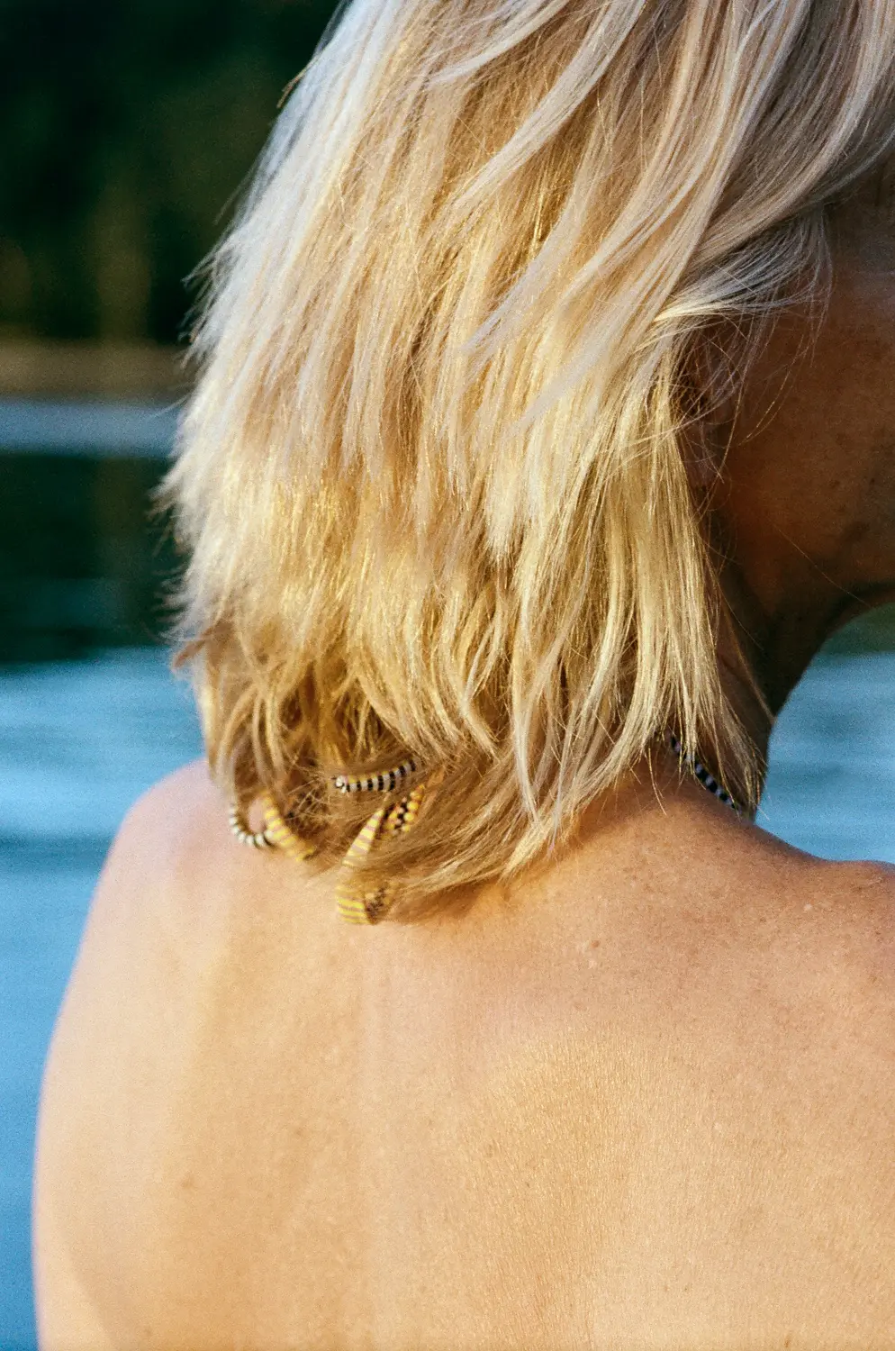 Das Bild zeigt einen Rücken einer Person mit blondem, gewelltem Haar. Die Person sitzt am Ufer eines ruhigen Sees. Eine Kette aus goldenen Federn hängt um den Hals der Person. Im Hintergrund sind Bäume und das Wasser des Sees zu sehen. Das Licht ist weich und diffus.