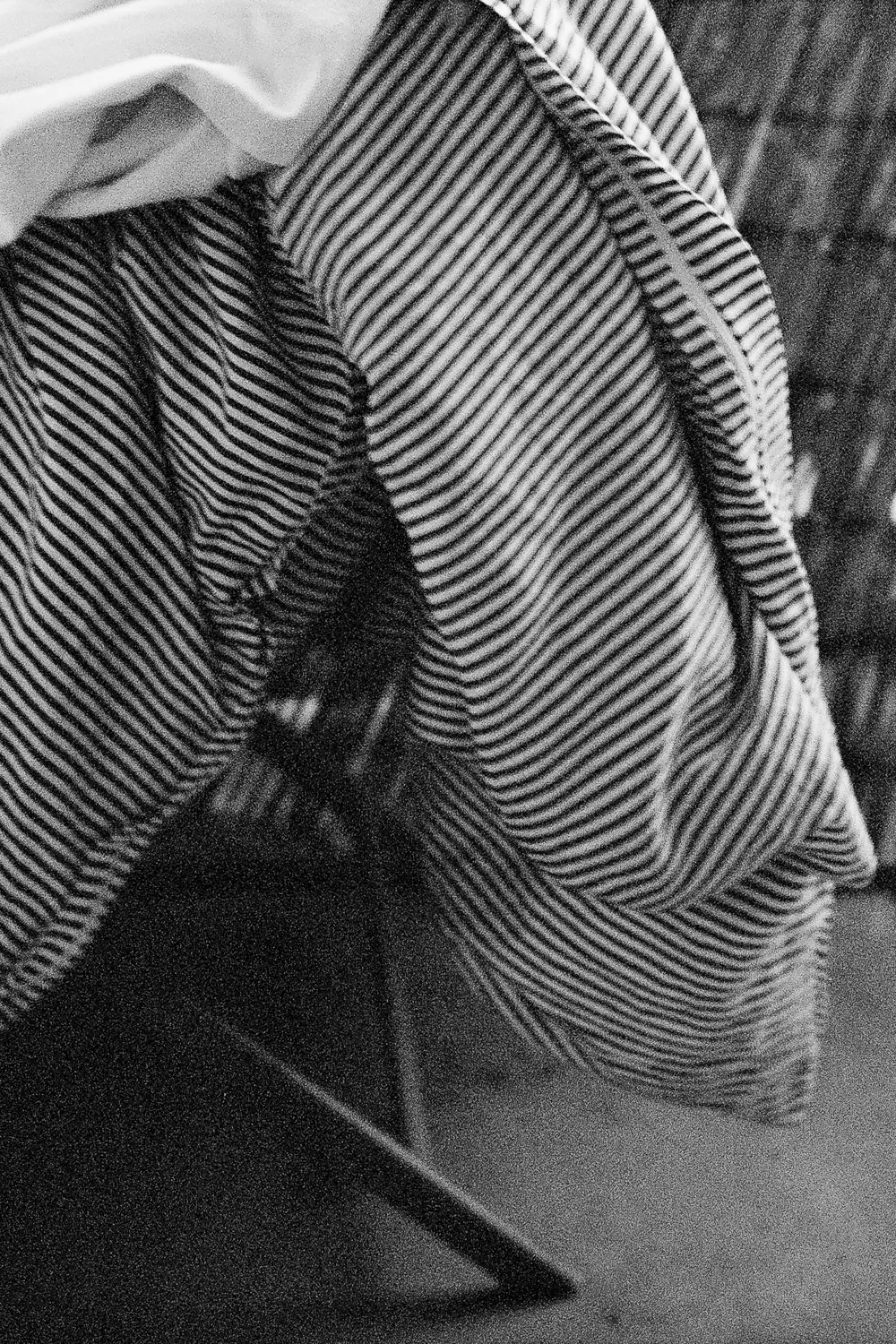 Die Schwarzweißfotografie zeigt eine Person, deren Gesicht durch ein gestreiftes Tuch verdeckt wird. Das Tuch ist diagonal über die Schultern gelegt und erzeugt dynamische Linien. Die Szene spielt sich im Freien ab, vermutlich auf einer Straße oder einem Gehweg. Der Hintergrund ist verschwommen und trägt zu einer düsteren Stimmung bei. Die Beleuchtung ist diffus und betont die Textur des Stoffes.