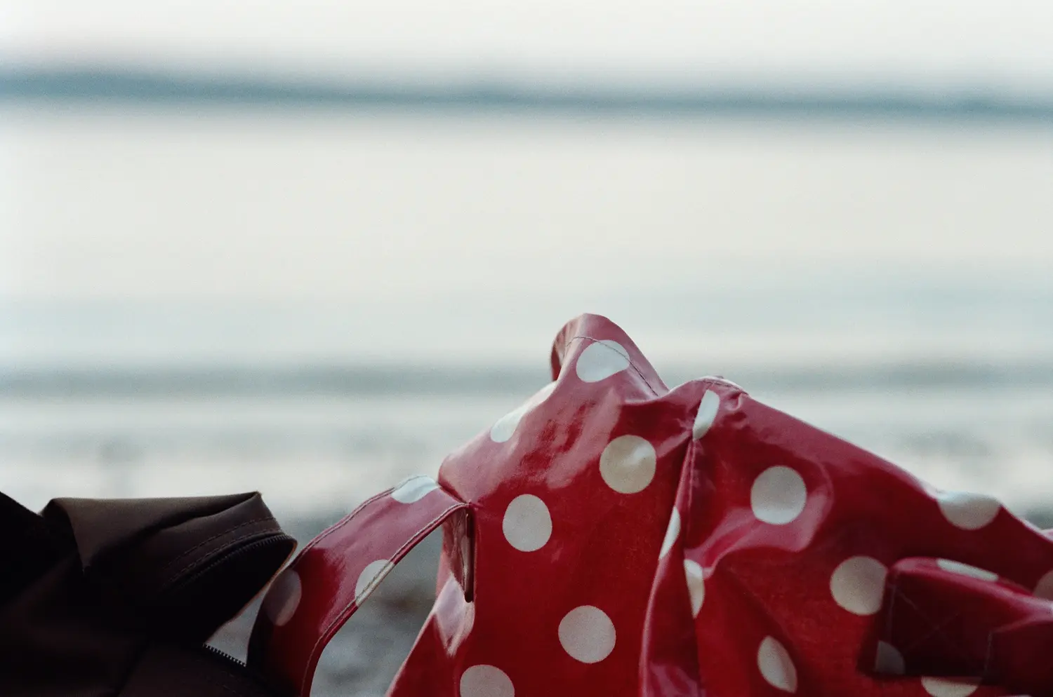 Die Aufnahme zeigt eine rote Strandtasche mit weißen Punkten, die am Ufer des Meeres liegt. Die Tasche ist leicht zerknittert und spiegelt das Licht des Wassers wider. Im Hintergrund erstreckt sich ein ruhiges, graues Meer. Der Fokus liegt auf der Tasche, wodurch die Details der Stoffmuster und der reflektierten Lichteffekte hervorgehoben werden.