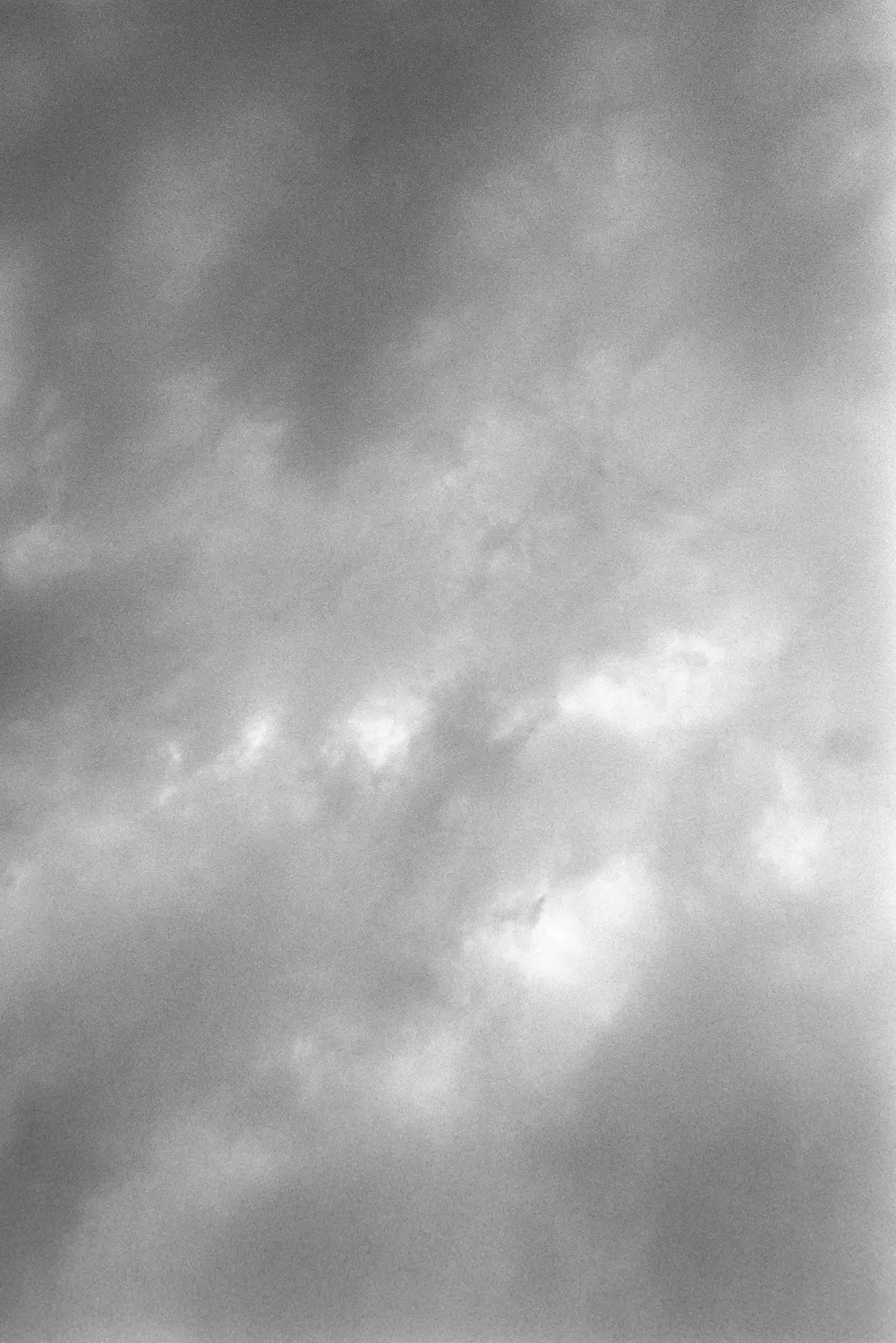 Die Aufnahme zeigt einen grauen Himmel mit vereinzelten weißen Wolken. Die Farben sind gedämpft und die Textur weist eine körnige Qualität auf, was auf eine ältere oder analoge Fotografie hindeutet. Es gibt keine erkennbaren Strukturen oder Objekte im Vordergrund.