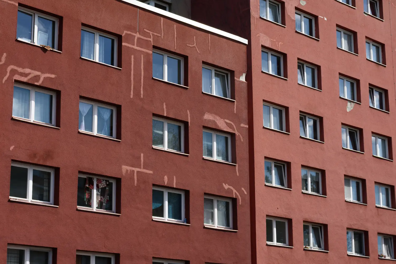 Die Aufnahme zeigt eine rote Wohnhausfassade, vermutlich im Stadtteil Berlin-Kreuzberg. Das Gebäude ist ein typisches Beispiel für den Wohnungsbau der DDR. Es besteht aus mehreren Geschossen und weist viele Fenster auf. Auf der Fassade sind Graffiti in Form von weißen Linien gemalt. Einige Fenster zeigen Stoffstücke oder Vorhänge. Die Beleuchtung ist hell, was die Details der Fassade hervorhebt.