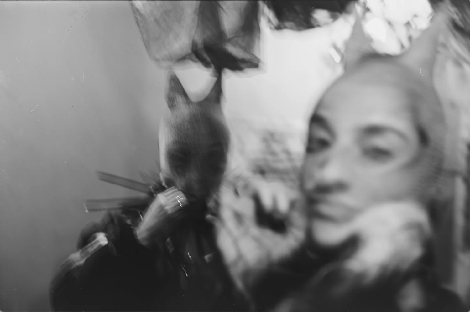 Das Schwarzweißfoto zeigt eine junge Frau, die ein Hasenkostüm trägt. Der Hase ist mit einer Maske versehen, die den Kopf des Hasen darstellt. Die Frau hat einen grauen Wollschal um den Hals und blickt direkt in die Kamera. Im Hintergrund sind verschwommene Elemente zu erkennen, was dem Bild eine surreale Atmosphäre verleiht. Das Licht ist diffus und erzeugt Schatten, die die Szene zusätzlich verstärken.