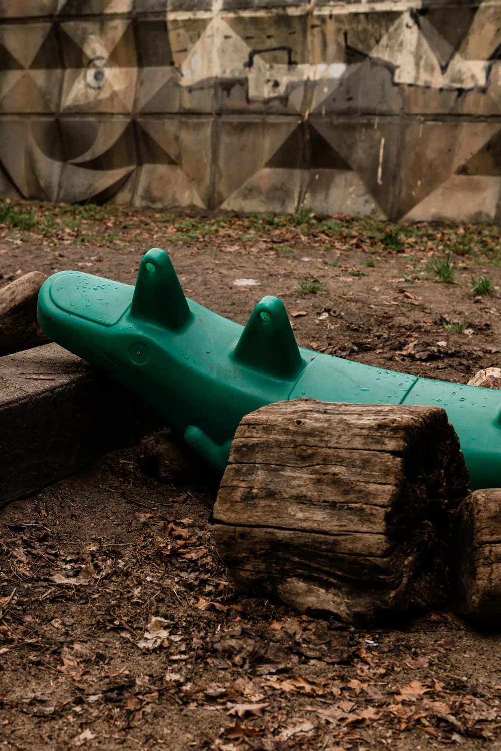 Die Aufnahme zeigt einen grünen Kunststoffschaukeldrachen, der auf einem Spielplatz in Berlin liegt. Der Drache ist leicht beschädigt und mit Wasser bedeckt, was auf Regen hindeutet. Im Hintergrund befindet sich eine Betonmauer mit einem geometrischen Muster. Die Szene ist düster und unruhig, mit einer starken Kontrast zwischen dem leuchtenden Grün des Drachen und den dunklen Schatten. Der Boden besteht aus Erde und Gras.