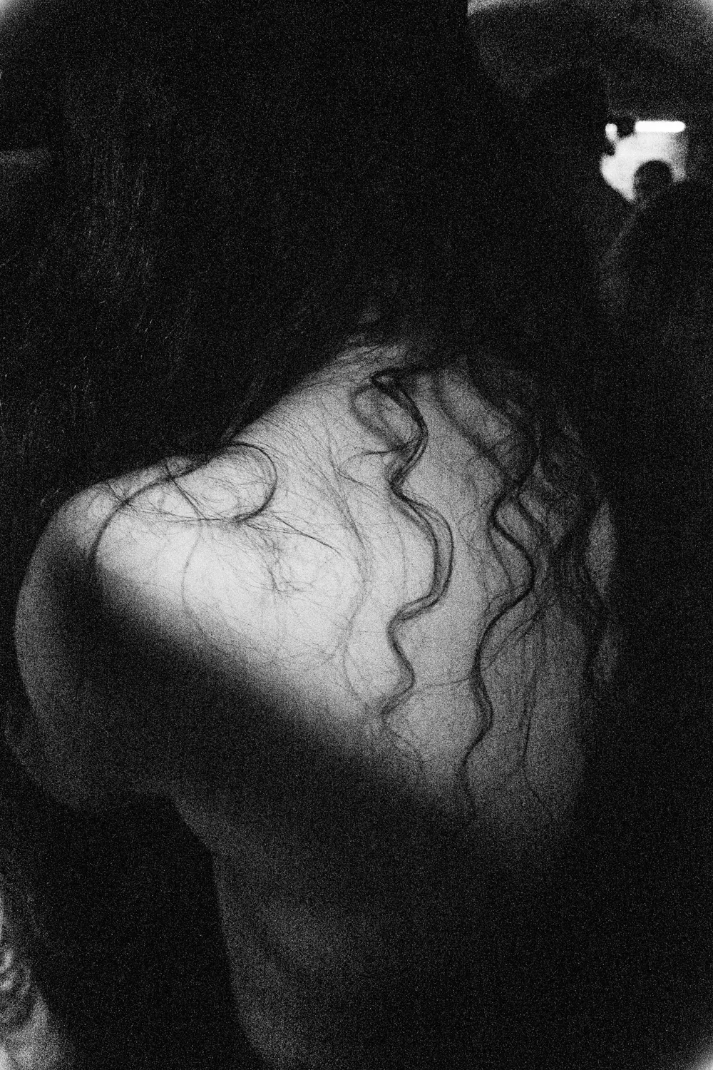 Die Schwarzweißfotografie zeigt einen menschlichen Schulterbereich im Vordergrund, wobei der Fokus auf dem Bereich um die Schulter liegt. Die Person hat lange, gewellte Haare, die über den Rücken fallen. Das Licht scheint von oben herab und erzeugt starke Schatten und Highlights, was die Textur der Haut und des Haares betont. Der Hintergrund ist dunkel und verschwommen, was die Aufmerksamkeit auf das Hauptmotiv lenkt.