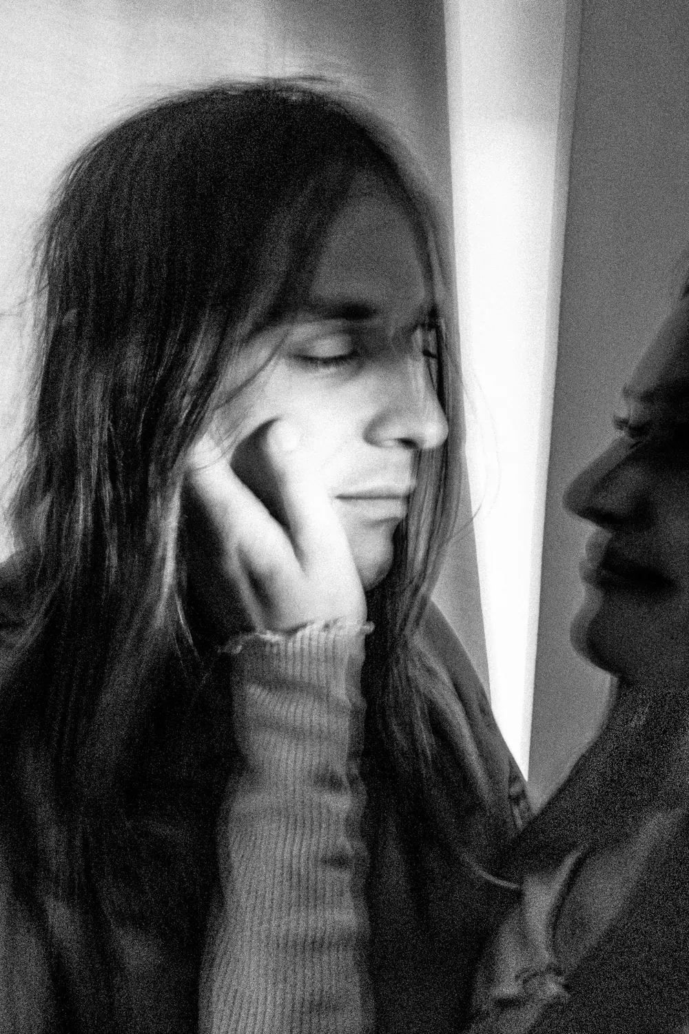 Dieses Schwarzweißporträt zeigt eine junge Frau mit langen, gewellten Haaren. Sie sitzt in einem Raum und scheint nachdenklich oder traurig zu sein. Ihre Hände sind an ihrem Gesicht, was auf eine innere Zerrissenheit oder ein Gefühl der Isolation hindeutet. Das Licht fällt durch das Fenster im Hintergrund und erzeugt einen dramatischen Effekt.