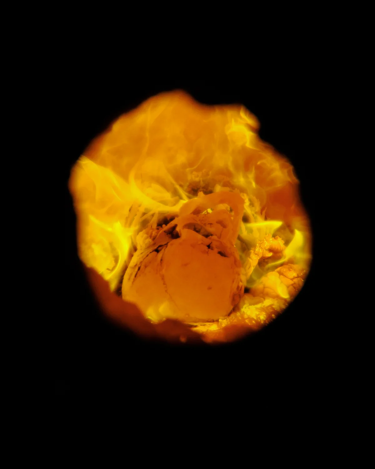 Die Aufnahme zeigt eine Kugel Eis, vermutlich Vanille oder Schokoladeneis, die vollständig von orangefarbenen bis gelblichen Flammen umgeben ist. Die Flammen sind dicht und erzeugen einen dramatischen Effekt. Der Hintergrund ist schwarz.